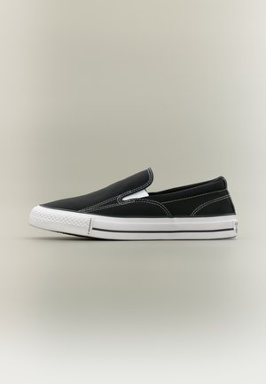 Svart slip-on canvas-sneaker med hvit gummisåle og hvit søm, plassert på siden på en nøytral bakgrunn.