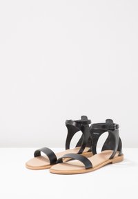 Musta nahkasandaalit, joissa on avonaiset varpaat, nilkkahihnat ja litteä pohja. Sisäpuolella on beigen värinen pinta kontrastiksi.