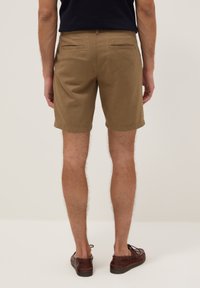 Hombre con pantalones cortos marrón claro hasta la rodilla, camisa negra y zapatos de cuero marrones, de pie de espaldas con fondo sencillo.