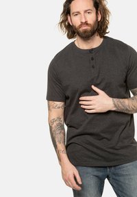 Graues Henley-Shirt mit kurzen Ärmeln, drei Knöpfen und rundem Ausschnitt. Standardpassform, glatte Textur, kombiniert mit blauen Jeans. Auffällige sichtbare Tattoos.