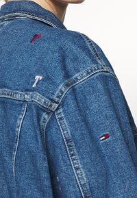 Spijkerjack met een blauwe tint, zichtbare stiksels en geborduurde accenten in rood en roze op de schouders. Klassiek kraagontwerp.