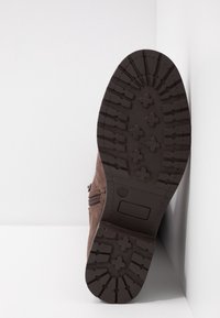 Semelle d'une botte marron avec des crampons profonds et des motifs en forme de croix, positionnée verticalement contre un coin blanc.