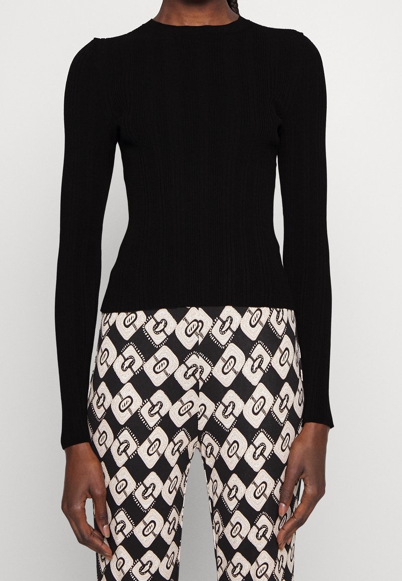 Personne portant un haut noir cintré à manches longues associé à un pantalon taille haute noir et blanc à motifs chaîne.