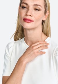 Balta trumpa rankove suapvalinta iškirpte ir subtiliomis pūstomis rankovėmis; lygus audinys su švelnia tekstūra; modelis liesia petį.