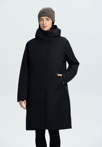 Cappotto nero alla ginocchia realizzato in materiale liscio e leggero, con cappuccio, tasche laterali e taglio dritto. Abbinato a un berretto di maglia marrone.