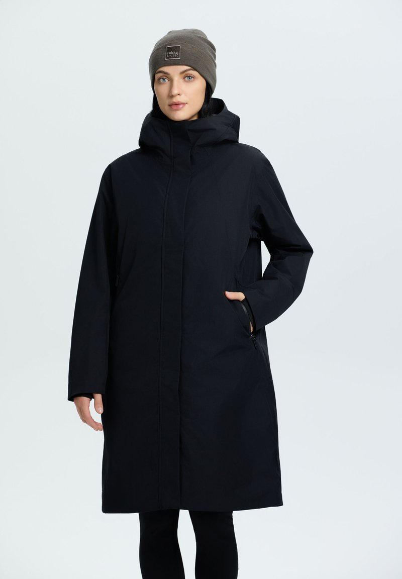 Cappotto nero alla ginocchia realizzato in materiale liscio e leggero, con cappuccio, tasche laterali e taglio dritto. Abbinato a un berretto di maglia marrone.