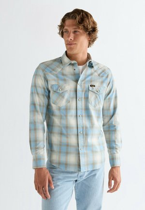 Homme portant une chemise à carreaux bleu clair et beige sur un t-shirt blanc, associée à un jean bleu clair, debout devant un fond blanc.