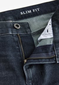 Jean slim bleu foncé avec fermeture éclair partiellement ouverte et bouton visible à la taille avec doublure de poche intérieure visible.