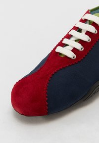 Zapato azul marino con una puntera de ante rojo, cordones blancos y bordes festoneados. Interior verde visible, con una suela texturizada.