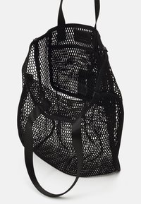 Svart mesh tote-väska med en semi-transparent design, utrustad med smala tyghandtag och en öppen topp. Insidan är texturerad och rymlig.