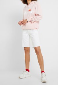 Sudadera rosa claro con logo blanco, pantalones cortos blancos desgastados y zapatillas blancas con acentos rojos. Tela suave, diseño informal.