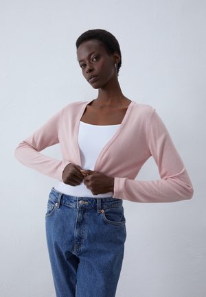 Cardigan rose clair à manches longues, décolleté en V et texture douce, porté sur un débardeur blanc et un jean bleu taille haute.