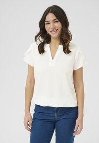 Blouse blanche à manches courtes avec un col en V et un tissu texturé, dotée de manches volantes et d'une coupe décontractée, associée à un jean bleu.