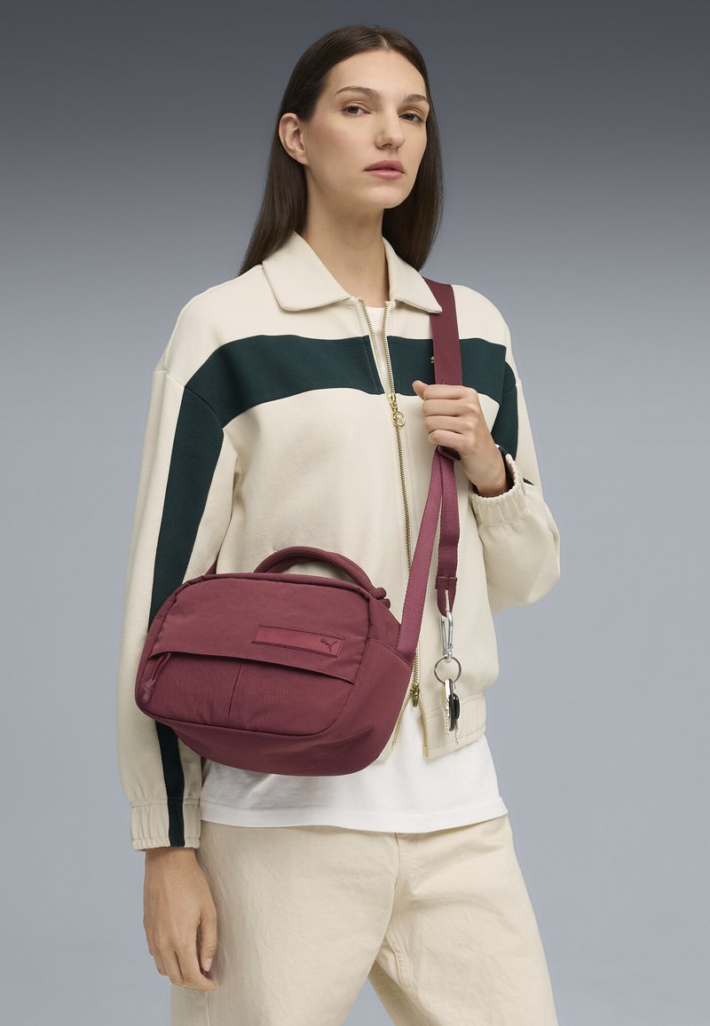 Borsa a tracolla bordeaux con finitura testurizzata, dotata di una tasca frontale e di una tracolla, tenuta da una persona in giacca avorio.