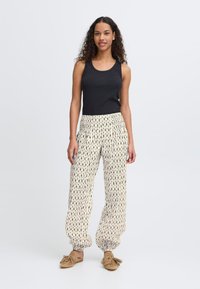 Débardeur noir côtelé associé à un pantalon à cordon crème à motifs. Le modèle porte des mocassins en suède beige. Fond uni.