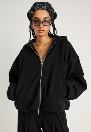 Felpa nera con zip, dal fitting rilassato, con vita elastica e un grande cappuccio. Il materiale sembra morbido e caldo, senza motivi o accenti visibili.