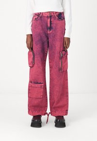 Pantalons cargo en denim rose avec une texture décolorée, présentant de grandes poches latérales, des fermetures éclair et des ourlets à cordon de serrage. Design à jambes larges.