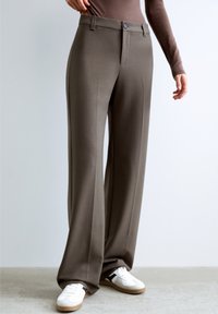 Wide-leg trousers in brown fabric with a smooth texture. Features a button closure and a classic design, paired with white sneakers.