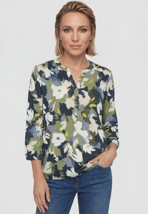 Donna con capelli biondi corti che indossa una camicetta a fantasia floreale con tonalità verdi, bianche, blu e gialle e jeans blu, mano in tasca.