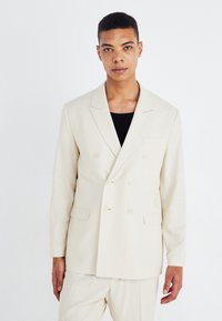 Blazer beige de doble botonadura hecho de tejido texturizado, con solapas de muesca, dos bolsillos frontales y un bolsillo en el pecho.