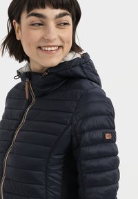 Navy puffer jacket with a hood, featuring a zip closure, quilted texture, and brown leather accents on the zipper and sleeve.
