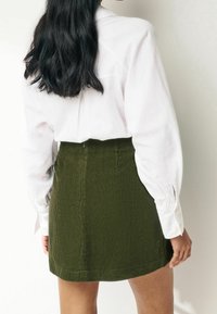 Weißes Langarmhemd mit geknöpften Manschetten, kombiniert mit einem olivgrünen Cord-Minirock mit strukturierten vertikalen Streifen.