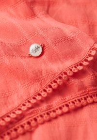 Tessuto corallo con una trama a quadri intrecciata, caratterizzato da una striscia decorativa in pizzo con pompon e un'etichetta circolare del marchio con il logo "Superdry".