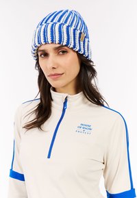 Gebreide beanie met blauwe en witte strepen, gemaakt van zacht garen. Het voorhoofdskanaal heeft een geweven label. Het model draagt een lichtbeige top met blauwe accenten.