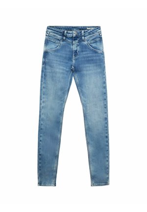 Obcisłe, wyblakłe niebieskie dżinsy noszone z czarnymi sandałami i białym krótkim topem, odsłaniającym brzuch i ręce luźno opadłe po bokach.