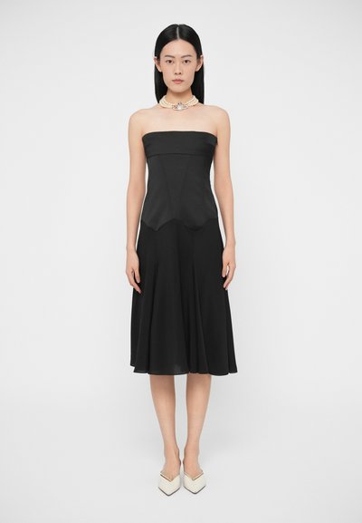 Mujer con un vestido midi negro sin tirantes, un collar choker de perlas y zapatos blancos de punta puntiaguda, de pie contra un fondo liso.