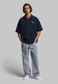 Granatowy oversize polo z krótkim rękawem i kołnierzykiem, z białym logo. Połączony z jasnoniebieskimi luźnymi dżinsami i białymi sneakersami.