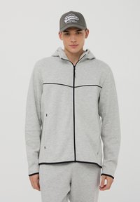 Felpa leggera grigia con zip e profili neri, dotata di cappuccio e tasche laterali, indossata con pantaloni grigi abbinati e un berretto grigio.