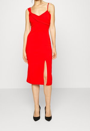 Vrouw draagt een nauwsluitende rode midi-jurk met een asymmetrische halslijn, schouderbandjes en een diepe split aan de voorkant van de dij, gecombineerd met zwarte puntige hakken.