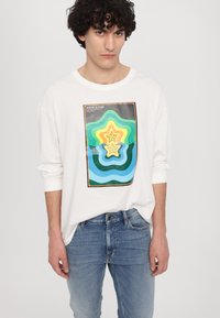 Hvit langermet t-skjorte med et fargerikt retro-stil grafikk med lagdelte former i grønne, blå og gule nyanser, kombinert med blå denim.