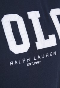 Marineblauwe stof met grote witte gestikte letters "OLD" boven kleinere witte tekst "RALPH LAUREN EST. 1967."