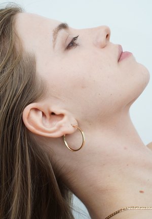 Goldener Creolenohrring an einem Model mit hellbraunen Haaren, mit einem glatten, abgerundeten Design und einem glänzenden Metallfinish.