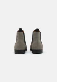 Par de botas Chelsea de ante gris con paneles laterales de elastano negros y lengüetas de tirón, vistas desde atrás sobre un fondo blanco.