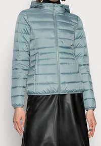 Světle modrá пуховка s zipem vpředu, kapucí a elastickými manžetami. Má horizontální prošívání a boční kapsy. Hladký, lesklý povrch.