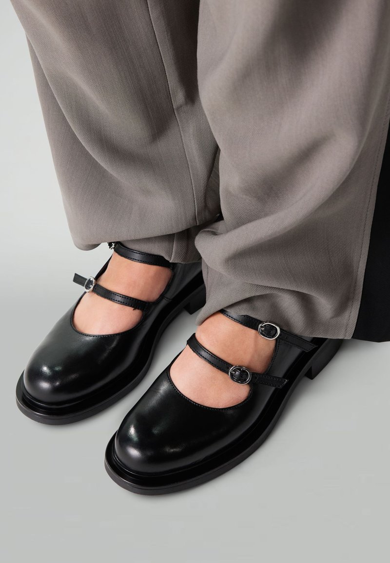 Chaussures Mary Jane en cuir noir avec deux brides ajustables, bout rond, finition laquée et semelle épaisse, portées avec un pantalon large gris.