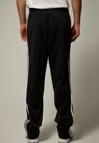 Czarne spodnie sportowe o luźnym kroju, z białymi paskami po bokach, elastycznym paskiem w talii oraz gładką, lekką teksturą materiału.