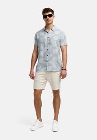 Hellblaues kurzärmliges Hemd mit weißen Blattmustern, dunklen Knöpfen, kombiniert mit beigen strukturierten Shorts und weißen Sneakers.