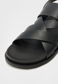 Svart lær sandaler med kryssede stropper, flat såle og teksturert overflate. Synlige sømmer og minimalistisk design med matt finish.