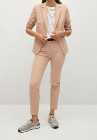 Costume rose clair avec blazer cintré et pantalon fuselé, associé à un t-shirt blanc et des baskets grises. Tissu lisse avec une texture minimale.