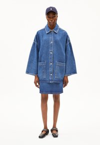 Denim jacket with large buttons, two front pockets, and wide sleeves. Paired with a denim skirt. Black shoes with studded accents. Navy cap.