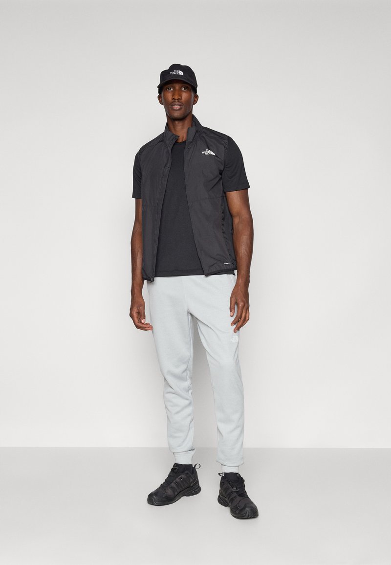 Un gilet sans manches noir sur un t-shirt noir, un pantalon de survêtement gris clair et des baskets noires. Le gilet arbore un logo sur la poitrine.