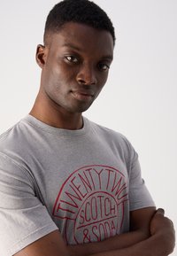Gray cotton t-shirt with a red circular logo featuring "TWENTY TWO SCOTCH & SODA." Slightly fitted design, short sleeves, and crew neck.
