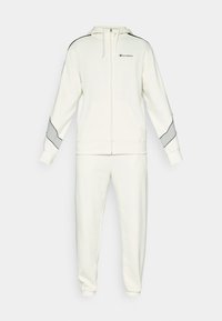 Sudadera blanca con cremallera y capucha, bolsillos frontales y rayas grises en los laterales. Combinada con pantalones de chándal ajustados con puños elásticos.