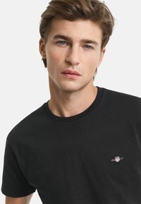 Schwarzes Baumwoll-T-Shirt mit Rundhalsausschnitt und kurzen Ärmeln. Ein kleines gesticktes Logo ist auf der linken Brust sichtbar.