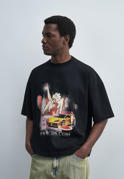 Man wearing black T-shirt with graphic of woman, yellow sports car, pasta plate, and website text, standing with hands in pockets.