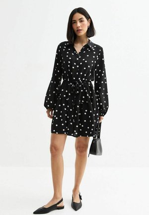 Black polka dot dress with long sleeves, cinched waist, and a flowing skirt. Features a button-up front and soft, smooth fabric. Worn with flats.